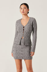 A woman is wearing a matching two-piece knit set consisting of a gray cable-knit cardigan with a deep V-neck and button closure, and a gray cable-knit skirt that is short and fitted. The knit fabric features an intricate diamond and cable pattern throughout.